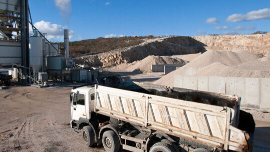 Dump truck at an asphalt plant with gravel piles and smoke-emitting processing tower under a clear sky.