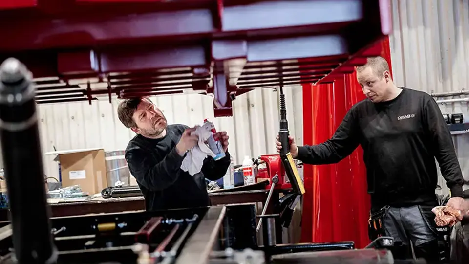 Two workers in black DISAB uniforms perform maintenance on a red industrial unit, using tools and cleaning supplies in a well-equipped workshop.