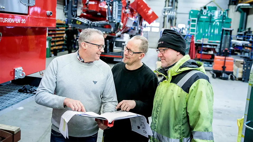 Three men discuss a manual in an industrial workshop, surrounded by DISAB vacuum equipment, with one wearing a high-visibility jacket.