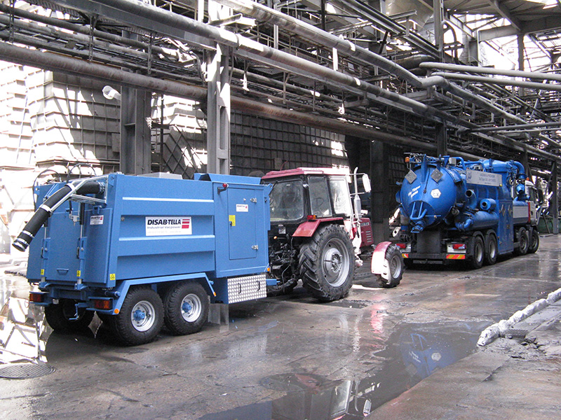 Blue DISAB TrailerVAC SDT-10T vacuum unit towed by a tractor through a wet, heavy industrial environment, demonstrating its mobility and use in raw material handling and site cleaning operations.