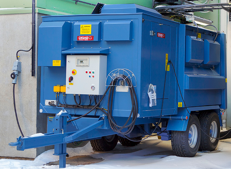 Blue DISAB TrailerVAC SET-10T industrial vacuum unit parked outside a building, showing the control cabinet, hose attachments, and hydraulic tailgate for efficient dust collection and safe material disposal.