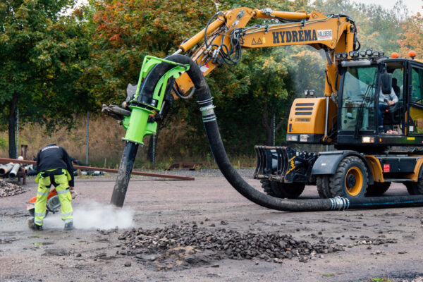 DISAB Hydrema vacuum excavator with bright‑green suction nozzle extracts gravel and dust, guided by a worker at an outdoor industrial site.