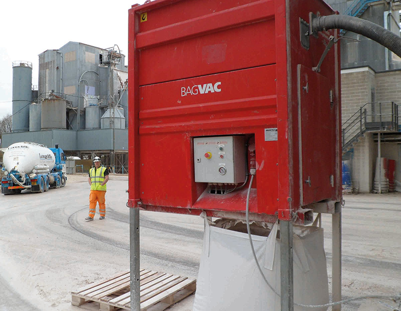 A red DISAB BagVAC industrial vacuum unit positioned outdoors on a factory site, with hoses attached and a worker in high-visibility clothing nearby, showcasing its mobility and use in heavy-duty industrial cleaning operations.