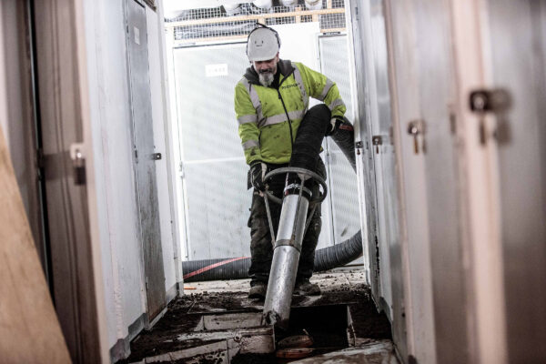 Operator in protective gear using a large suction hose from a DISAB unit inside a building, emphasizing safety, ergonomics, and material handling in confined industrial spaces.